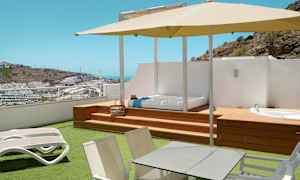 Terrasse med solsenger, bord, parasoll og utsikt over et kupert område med bygninger. (AI-generert tekst)