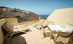 Terrasse med solsenger og bord med stoler, utsikt mot havet. (AI-generert tekst)