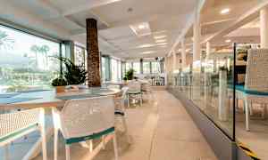 Interiøret i en restaurant med hvite stoler og bord, glassvegger og utsikt mot utendørs grøntområder. (AI-generert tekst)