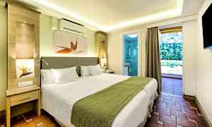 Et moderne hotellrom med to enkeltsenger, grønt sengeteppe og hvit innredning, med utsikt mot en balkong og bad. (AI-generert tekst)