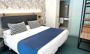 Et moderne hotellrom med dobbeltseng, blå sengeteppe og to mønstrede puter. (AI-generert tekst)