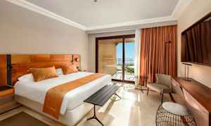 Et hotellrom med en stor seng, oransje interiørdetaljer, et skrivebord og en balkong med havutsikt. (AI-generert tekst)