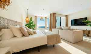 Bilde av et lyst og moderne soverom med stor seng, sofa, TV og en bred havutsikt gjennom store vinduer. (AI-generert tekst)