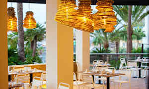 Utemøbler ved restaurant med hengende lampeskjerm av tre og palmetrær i bakgrunnen. (AI-generert tekst)