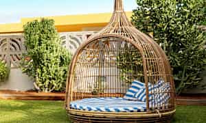 Et rottingmøbel i form av en baldakin med stripete blå og hvite puter står på en plen ved siden av noen grønne busker. (AI-generert tekst)