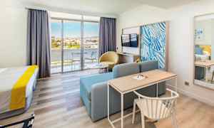 Et hotellrom med seng, skrivebord, sofa, TV og glassdører som fører til en balkong. (AI-generert tekst)