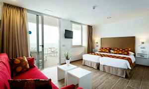 Et moderne hotellrom med to enkeltsenger, røde sofaer og en balkong med havutsikt. (AI-generert tekst)