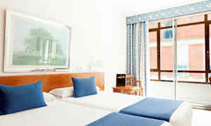 Et hotellrom med to enkeltsenger som har blåe tepper og puter, en stol ved et bord samt et vindu med blått gardin. (AI-generert tekst)