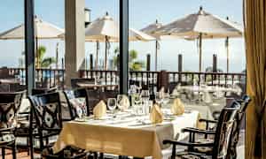 Et dekket bord i en restaurant med utsikt over en terrasse med parasoller og havet. (AI-generert tekst)