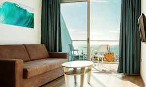 En stue med sofa og bord, med en balkong og utsikt over havet. (AI-generert tekst)