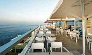 Bilde av en utendørsterrasse med bord, stoler og parasoller, med utsikt over havet. (AI-generert tekst)