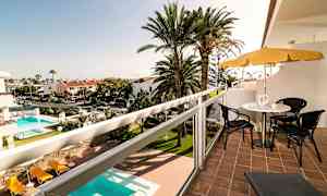 Utsikt fra en balkong med sittemøbler og parasoll, med et basseng og palmetrær i bakgrunnen. (AI-generert tekst)