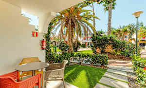 En terrasse med bord og fargerike stoler, rammet inn av en bue som fører ut til en hage med palmer og bygninger i bakgrunnen. (AI-generert tekst)