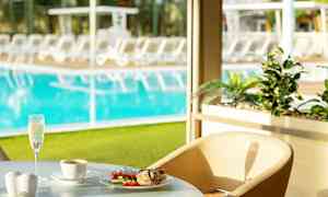 Et bord ved et vindu med en tallerken med mat, en kopp kaffe og et glass musserende drikke. Bassengområdet med solsenger kan ses i bakgrunnen. (AI-generert tekst)