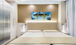 Bildet viser et moderne hotellrom med en dobbeltseng, stilrene sidebord og en veggdekor med tre bilder av et tropisk strandmotiv. (AI-generert tekst)