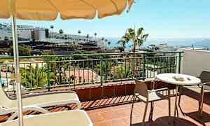 En terrasse med bord, stoler og solstoler. I bakgrunnen sees palmer, bygninger og havet. (AI-generert tekst)