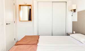 Et hotellrom med hvit innredning, en dobbeltseng med hvite laken og brune sengetepper, samt et speil på veggen. (AI-generert tekst)