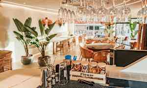 En moderne kafé med glass, store grønne planter og stoler i flettverk. (AI-generert tekst)