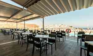 Uteservering med bord og stoler, utsikt over by og hav. (AI-generert tekst)