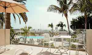 En balkong med solsenger og et bord med stoler, utsikt over et basseng og palmer. (AI-generert tekst)