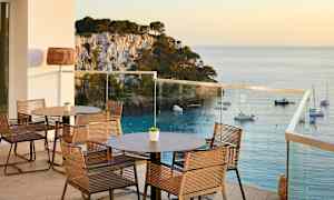 En terrasse med bord og stoler, utsikt over havet og båter ved en steinete kyst. (AI-generert tekst)