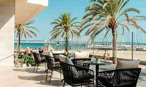 Uteservering med sitteplasser og bord med sjøutsikt, palmer i bakgrunnen. (AI-generert tekst)