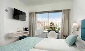 Et hotellrom med to senger oppredd med lyse laken og turkise sengetepp. En stol står foran en glassdør som leder ut til en balkong. En veggmontert TV henger ved siden av en hylle med vannflasker og kaffemaskin. (AI-generert tekst)