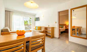 En lys stue med spisebord, stoler, sofa og balkong med utsikt over grøntområder. Et tilstøtende soverom er også synlig. (AI-generert tekst)