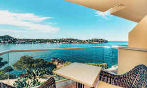 Utsikt fra en balkong mot en vik med klart blått vann, omgitt av kupert terreng og trær. Et bord og stoler vises i forgrunnen på balkongen. (AI-generert tekst)