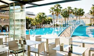 Et restaurantområde med uteservering ved et basseng omgitt av palmer og bygninger. En blå gangbro strekker seg over bassenget. (AI-generert tekst)