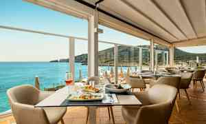 En restaurant med bord med mat og stoler, samt en panoramautsikt over havet og en fjellkyst. (AI-generert tekst)