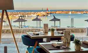 Utsikt fra en restaurant mot en strand med parasoller og solsenger, samt en småbåthavn med båter. (AI-generert tekst)