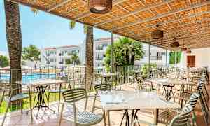 En utendørsrestaurant med bord og stoler under et tak, med utsikt over et basseng og hotellbygninger i bakgrunnen. (AI-generert tekst)