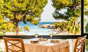 Et dekket bord med mat og to glass vin, med utsikt mot sjøen gjennom et stort vindu. (AI-generert tekst)