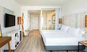 Et lyst hotellrom med trematerialer og moderne møbler. Rommet har en stor seng, en TV og en åpen løsning. (AI-generert tekst)