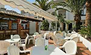 En uteservering med hvite stoler og bord under en markise, omgitt av palmer og grøntområder. (AI-generert tekst)