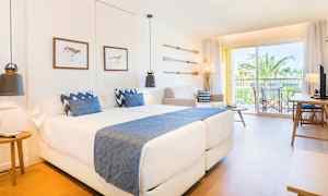 Et moderne hotellrom med to senger og blå detaljer samt en balkong med utsikt mot palmer. (AI-generert tekst)