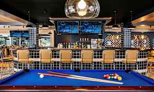 Biljardbord med blå duk foran en bar med stoler og TV-skjermer. (AI-generert tekst)