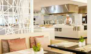 Et moderne restaurantområde med hvite møbler, pastellfargede puter og planter på bordene, i bakgrunnen er et åpent kjøkken synlig. (AI-generert tekst)