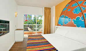 Et hotellrom med en dobbeltseng og fargerikt sengeteppe, et mønstret veggmaleri bak sengen, et vindu med utsikt over balkong og grøntområder. (AI-generert tekst)