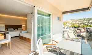 Et hotellrom med to senger og en sittemøbel, med en tilknyttet balkong hvor det finnes bord og stoler samt utsikt over en frodig ås. (AI-generert tekst)