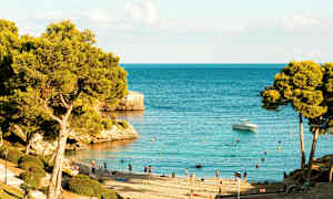 Strand med badegjester og trær ved et klart blått hav, en båt er synlig på vannet. (AI-generert tekst)