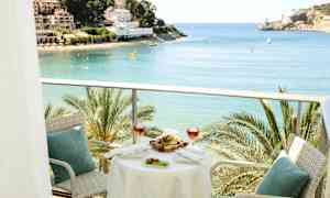 En balkong med bord og stoler, med utsikt over en bukt med palmer og bygninger i bakgrunnen. (AI-generert tekst)