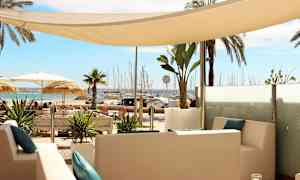 Utsikt over en rolig sittegruppe med hvite møbler og blå puter, omgitt av tropiske planter, med en strand og en småbåthavn i bakgrunnen. (AI-generert tekst)