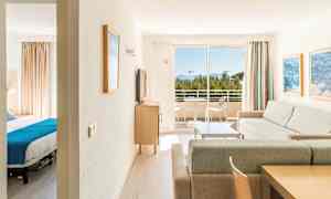 En lys hotellsuite med en stue, et soverom og en balkong som har utsikt over trær og hav. (AI-generert tekst)