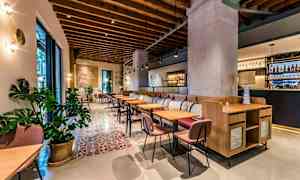 Et stilrent restaurantmiljø med tretakdetaljer, komfortable stoler og bord, samt dekorative planter. Et barområde er synlig i bakgrunnen. (AI-generert tekst)