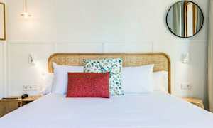 Et hotellrom med en oppredd dobbeltseng, pyntet med hvite puter og en rød dekorasjonspute foran en med blomstrete mønster. Sengegavlen er av flettet tre. Et rundt speil og vegglamper vises på en lys bakgrunn. (AI-generert tekst)