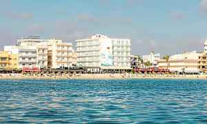 Utsikt fra havet mot en strand med flere bygninger og mennesker som soler seg. (AI-generert tekst)