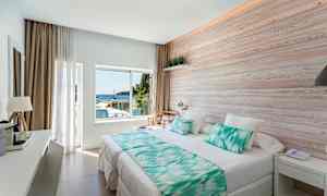 Et lys hotellrom med en stor seng, trepanel på veggen og et vindu som vender mot havet. (AI-generert tekst)