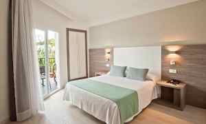 Et hotellrom med en dobbeltseng, to puter og et grønt teppe. Sengen er plassert nær en balkong med glassdører. (AI-generert tekst)